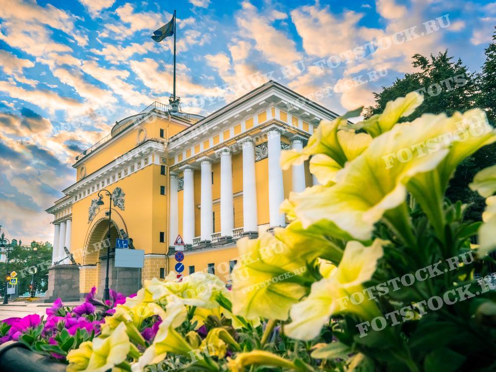На фотографии запечатлено Адмиралтейство в Санкт-Петербурге — один из главных символов города. На переднем плане видны красочные цветы, добавляющие живописности кадру, а на заднем плане — величественное здание с колоннадой и флагом на крыше. Над всем этим возвышается яркое небо с облаками. Фото может использоваться в туристических и путеводительных материалах, в образовательных целях для знакомства с архитектурой Санкт-Петербурга или как элемент декора, отражающий культурное наследие города.
